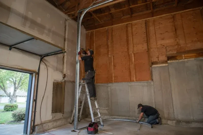 One person on a stepladder working beside a tall curved garage-door track while another kneels beside loose track sections in an unfinished garage. — High-Lift Garage Door Conversion, King